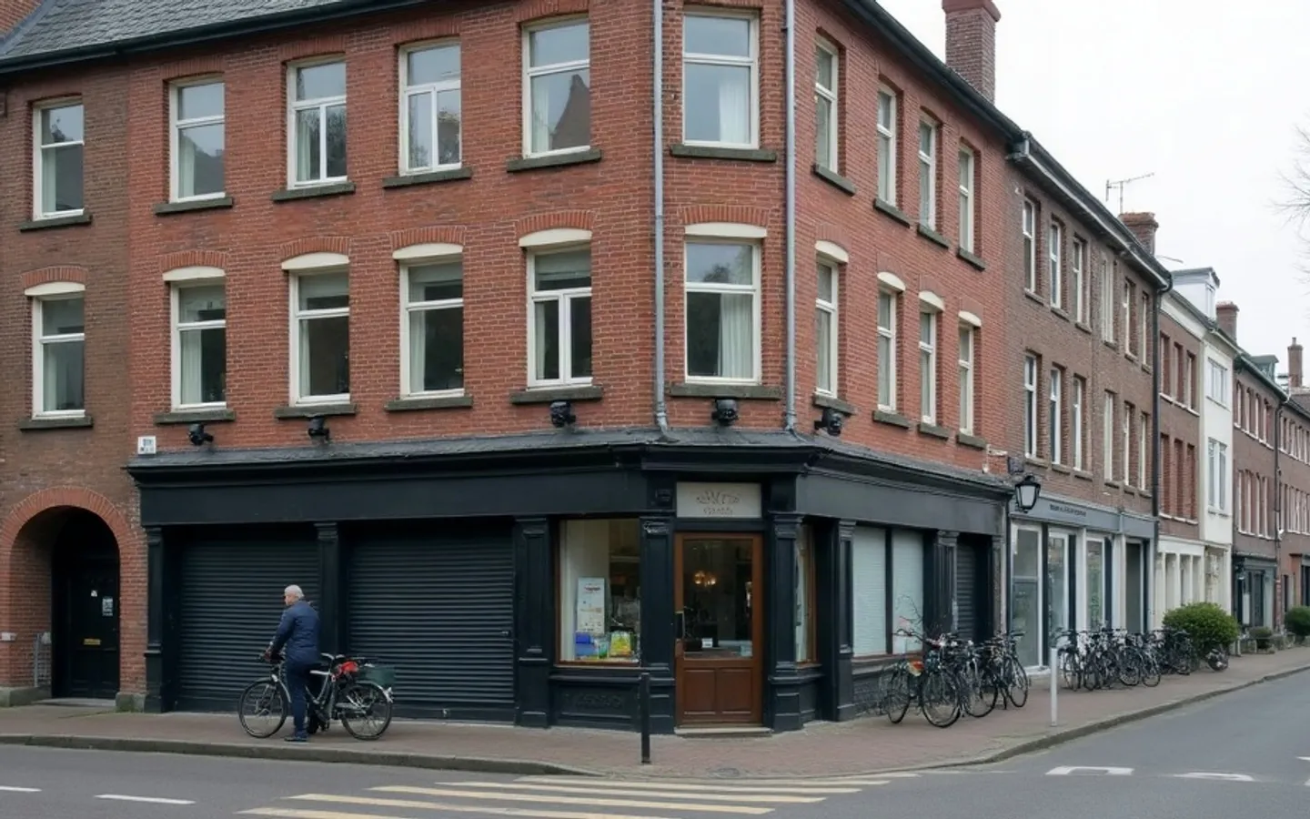 Een Nederlands hoekpand met een winkel op de begane grond en twee woningen erboven, gefotografeerd vanaf de overkant van de straat in het ochtendlicht. Een fiets staat tegen de bakstenen gevel.