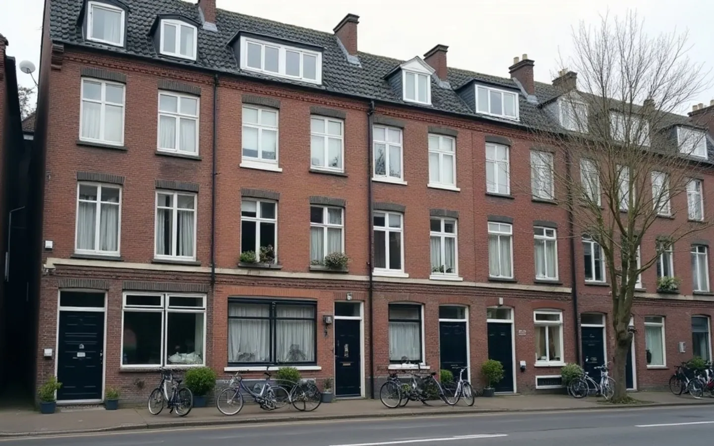 Een rij bakstenen appartementengebouwen aan een rustige stadsstraat in het ochtendlicht, met fietsen bij de ingangen en gordijnen voor enkele ramen.